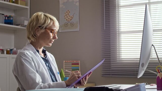 Side view waist up of mid aged Caucasian female pediatrician writing on clipboard and talking while looking at computer in clinic office. Ideal for healthcare services and pediatric clinic marketing