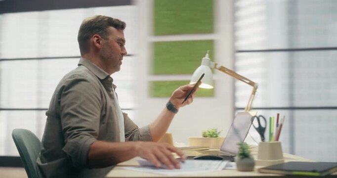 360 Degree. Man Using Smartphone While Working in Modern Loft Office, Scrolling E-commerce App and Social Media Feed and Reviewing Sales Data on Laptop and Printed Reports. Orbital Arc Tracking Shot.