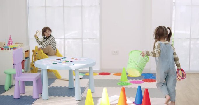 Asian female preschool teacher watches toddler girl carry plastic basket, toddler boy waits near play table, cone obstacle course in daycare room, ring toss game, slow motion, fun learning skill