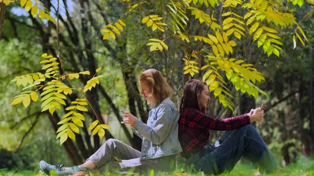 Two young women, a blonde and a brunette, are sitting back to back on a lawn among autumnal trees and using their smartphones. A loving LGBT couple enjoys a sunny autumn day in the park.
