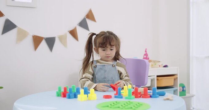 Asian toddler girl playing peg stack set alone on round table, colorful pieces arranged, daycare classroom bunting decor, independent learning, concentration, slow motion childhood education concept.