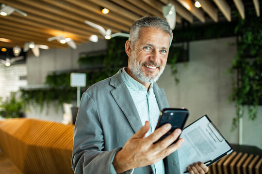 Smiling mature businessman using smartphone in modern office lobby