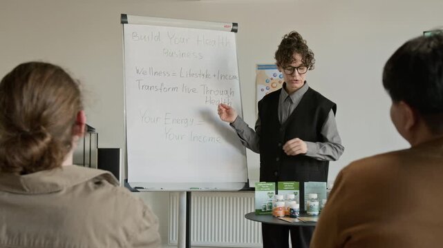 Young woman with glasses presenting health business strategy on whiteboard and showing supplement bottle to diverse audience. Clip for wellness coaching and marketing campaigns