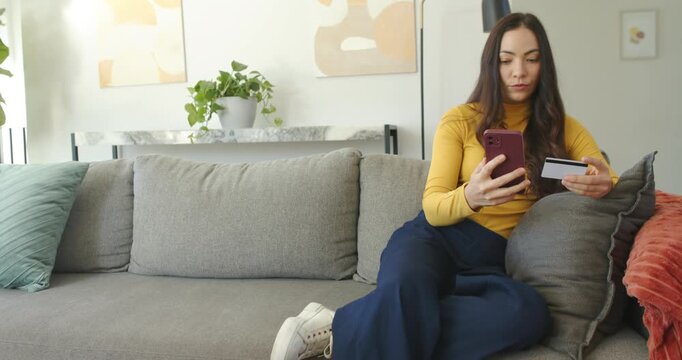 Woman wearing mustard top sitting on sofa with card, holding phone, scrolling, paying, copy space