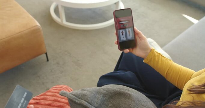 Woman in mustard top browsing clothing on smartphone, retrieving credit card on sofa for payment