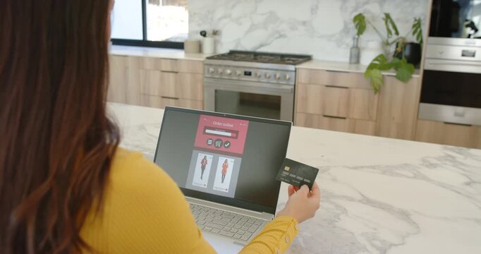 Woman viewing checkout UI, holding credit card and typing to pay at kitchen counter