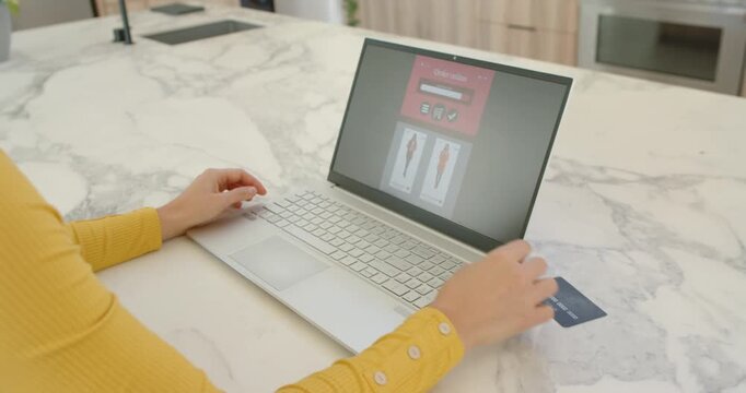 Woman in yellow top at kitchen island finishing laptop checkout, typing, holding credit card to pay