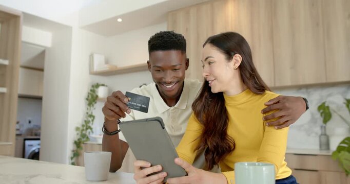 Diverse couple sitting at kitchen counter, man offering card, woman tapping tablet, paying