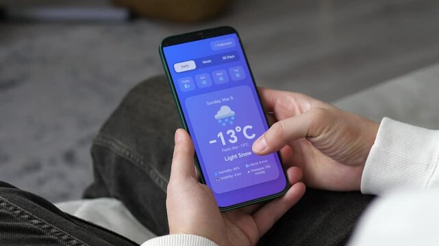 Viewing a daily weather forecast on a smartphone. The screen displays a temperature of minus 13 degree with light snow and a blue UI. Perfect for weather apps, climate visuals, and daily lifestyle