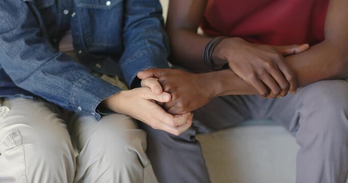 Couple sitting on couch, woman in denim placing hands around man's hand with bracelets comforting