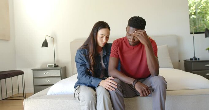 Diverse couple sitting on bed woman talking touching knee leaning in calming man after head bowing