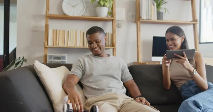 African American couple laughing, tapping tablet and discussing topic on couch at home with headset
