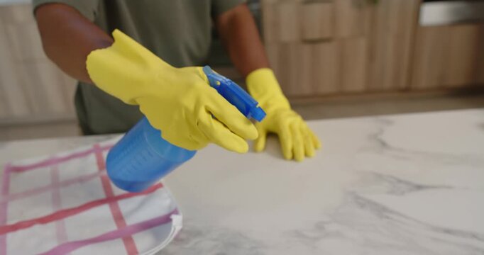 Adult woman in yellow gloves squeezing blue sprayer spraying on marble top in kitchen cleaning