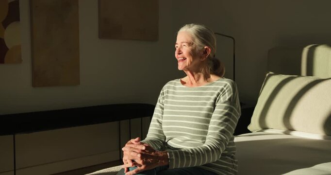 Senior woman sitting on bed in striped top, sunlight beaming right guiding head turning and smiling