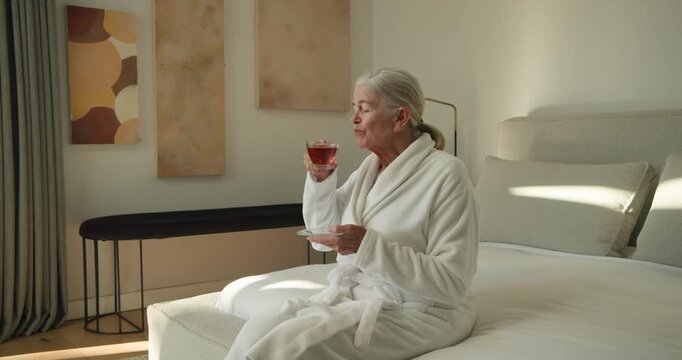 Senior woman sitting on sunlit bed in robe holding glass teacup on saucer sipping, seeking calm