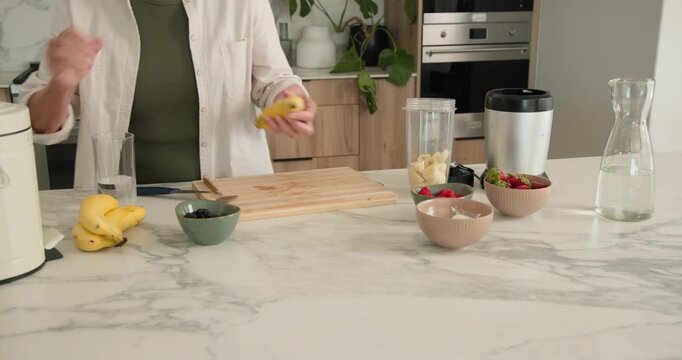 Senior woman placing banana in blender peeling and slicing on board marble counter making smoothie