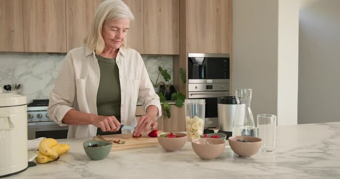 Senior woman wearing green top slicing strawberries on board for blender cup at kitchen island