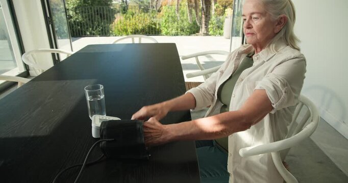 Senior woman sliding arm into cuff pressing monitor control checking BP at dining table, copy space
