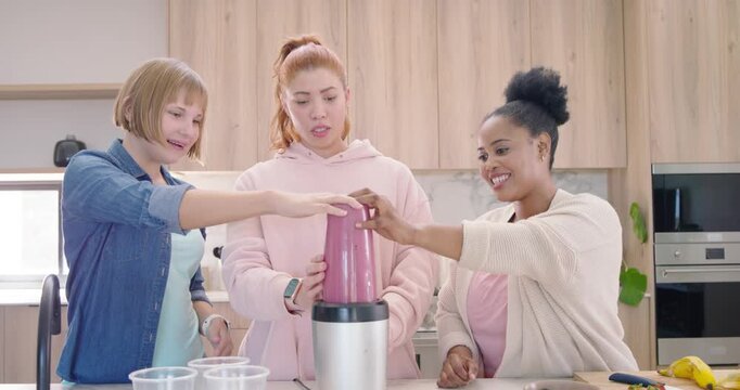 Diverse female friends making smoothie placing pink cup on base at counter, steadying, aligning