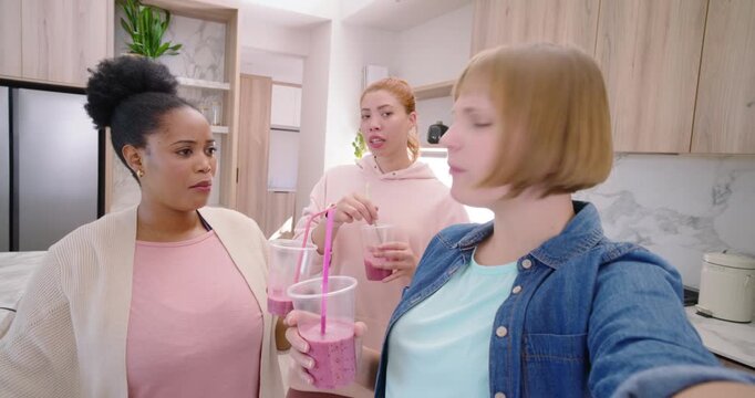 3 women in kitchen filming selfie for fun as right woman raising phone, sipping pink drinks