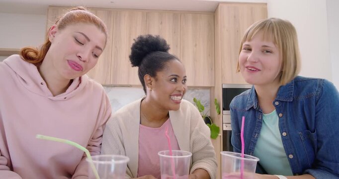 Female friends after fingertip covering lens, perking up, tasting pink smoothies at kitchen counter