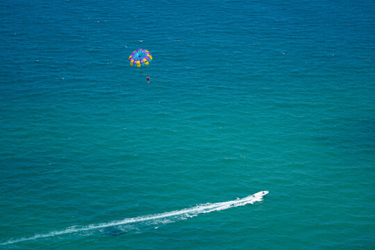 A person experiences parasailing, soaring high under a colorful parachute above the clear turquoise ocean