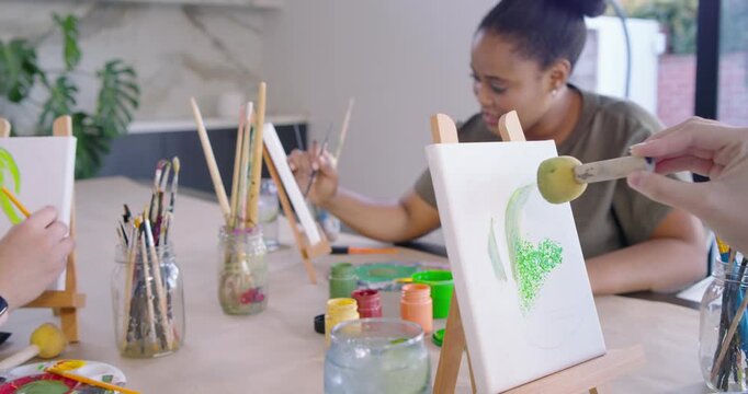 Diverse women painting at table, one in olive top dipping sponge into green paint, stamping canvas