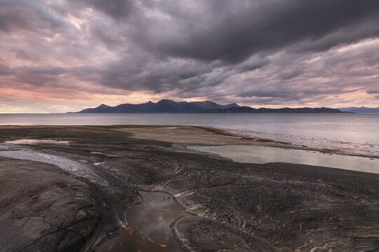 The Vestfjord in Nordland, Norway, offers a dramatic atmosphere near Bod&oslash;. Dark clouds are illuminated by the setting sun in shades of red and yellow. The rugged cliffs and mountains are reflected in the quiet fjord, which offers a breathtaking view of Lofoten