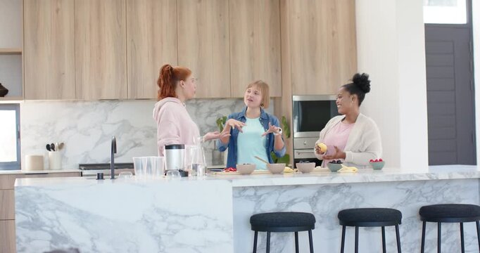 Diverse women chatting while prepping fruit on marble island in kitchen, tossing banana for laughs