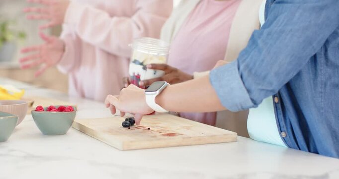 Three women making yogurt parfait at kitchen counter after cutting berries with knife for snack