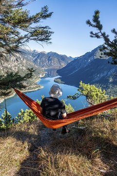Young man sitting in red hammock with panoramic view of mountains and lake, Plansee, Ammergau Alps, Reutte district, Tyrol, Austria