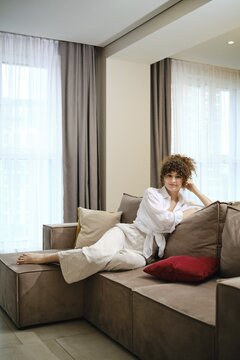 A woman with curly hair is lounging comfortably on a plush sofa in a modern living room. Soft natural light filters through the curtains, creating a calming atmosphere