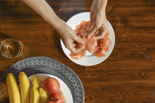 Fresh ingredients are being carefully arranged on a wooden table. A person is separating pieces of salmon on a plate, while bananas and apples are nearby, emphasizing a healthy eating lifestyle
