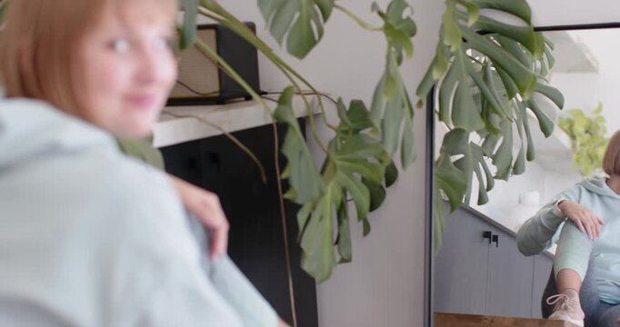 Woman adjusting hair while camera framing mirror and Monstera at home checking look, smiling at cam