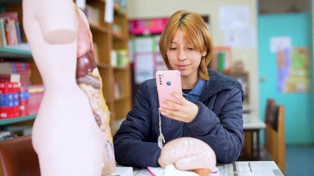Young woman studying human anatomy with a mobile phone in a school library next to anatomical models