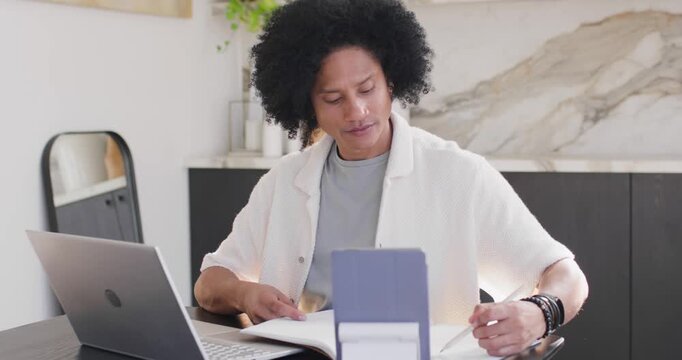 African American man at home office checking tablet, flipping binder and typing report on laptop