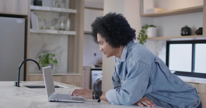 African American man in denim leaning over marble island noticing laptop screen, typing for work