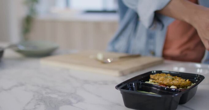 After plating, African American man in denim sealing trays with clear lids at counter for storage
