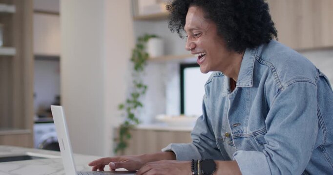African American man reacting to message, smiling, laughing and typing on laptop at kitchen island