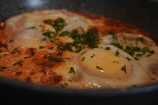 Shakshuka Eggs Cooking in Tomato Sauce with Fresh Parsley Garnish in Dark Frying Pan Overhead View for Breakfast Brunch Cuisine