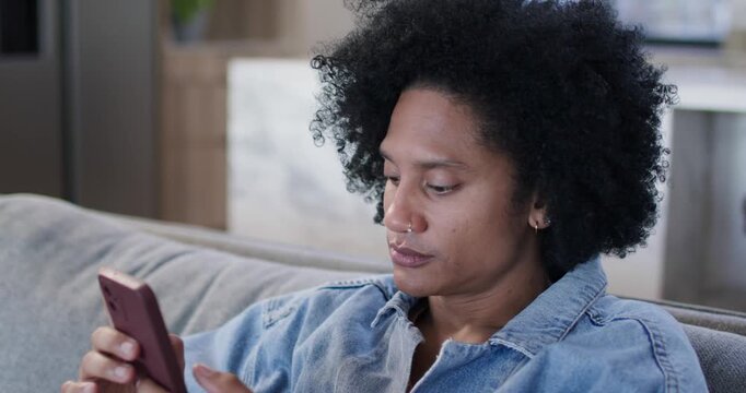 African American woman in denim jacket scrolling mauve phone on sofa at home, changing to bemused