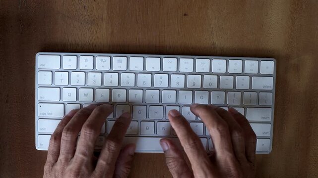 Close up top down view of Asian male finger busy typing on modern wireless computer keyboard with ten finger on wooden table