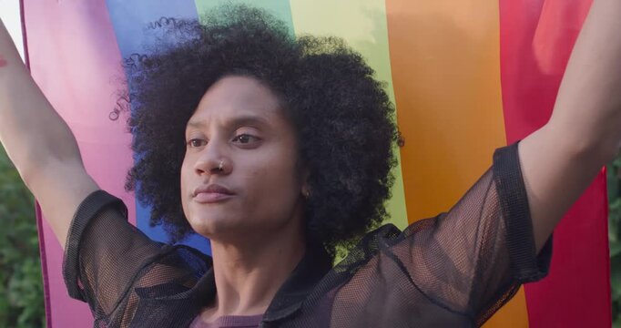 Adult African American man starting by holding rainbow flag, raising arms, showing pride in yard