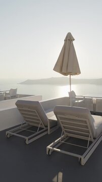 White architecture in Santorini island, Greece. Terrace with chaise lounges and sun umbrella. Beautiful sea view at foggy sunset. Travel and summer vacation concept. Vertical video
