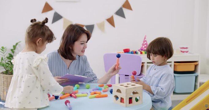 Asian female daycare teacher smiling, holding clipboard, observing toddler boy, toddler girl building magnetic construction set on table, education assessment, creativity, teamwork, STEM lesson.