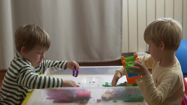 Siblings engage together over shared sensory table with pink and green kinetic sand capturing social bonding and parallel play that supports early childhood cooperation skills. 