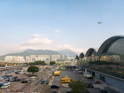 Hong Kong, China - June 19, 2026: A panoramic view of Hong Kong International Airport showcasing modern architecture, parked vehicles, and a distant airplane flying above. The scene is set against lus