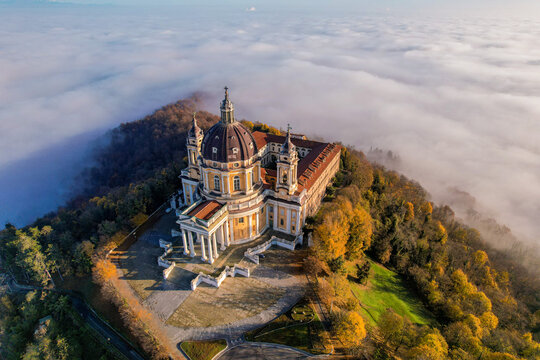 Aerial view of the Basilica of Superga standing above a thick layer of fog with autumn trees on the hillside in Superga, Piemonte, Italy.