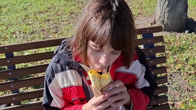 Child enjoys snack on bench in park while sitting under a tree during sunny day