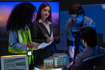 Fototapeta premium South Asian male construction professional presents architectural plans while colleagues review a scale model in a dark smart building control room.